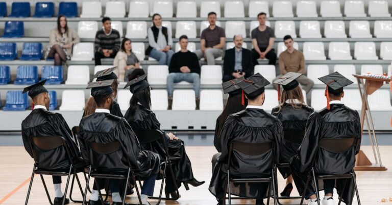 Graduates in gowns sit in a gymnasium with an audience in the bleachers, capturing a special moment.