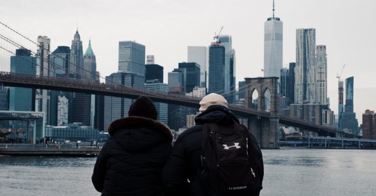 Two people enjoy the iconic view of the Manhattan skyline and Brooklyn Bridge from the riverbank.