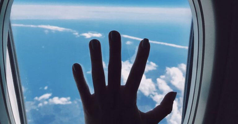 Silhouette of a hand touching an airplane window with a view of clouds and the sky.