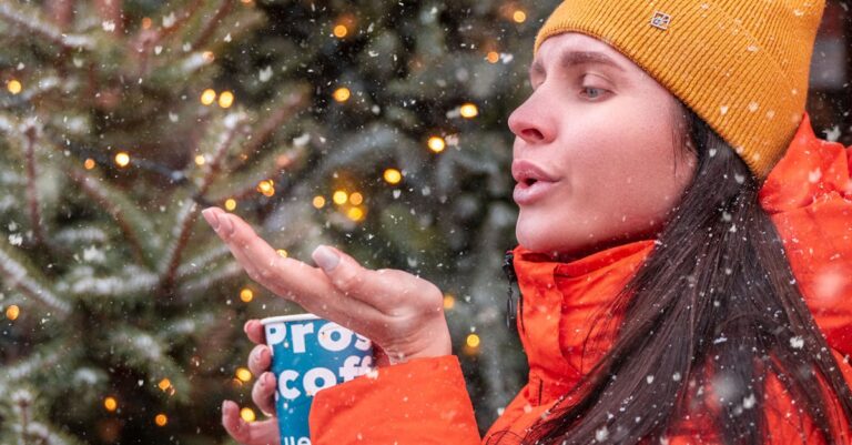 Woman wearing beanie and jacket in snow, enjoying a hot drink outdoors.