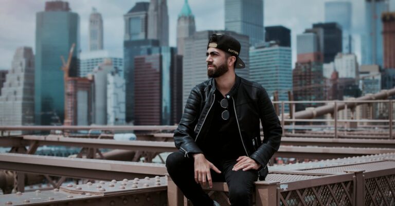 A man wearing a black leather jacket sitting on Brooklyn Bridge with New York City skyline in the background.