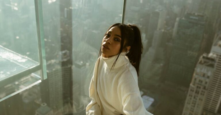 Young woman in cozy sweater posing by a glass wall overlooking New York City from a skyscraper.