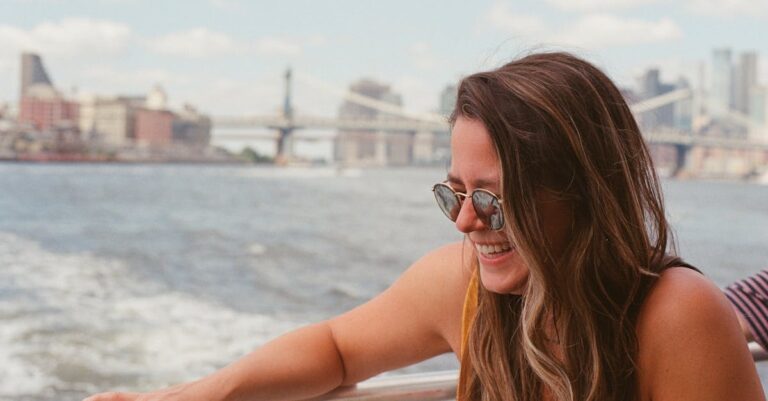 Smiling woman in sunglasses enjoys a summertime boat ride with New York City skyline in the background.