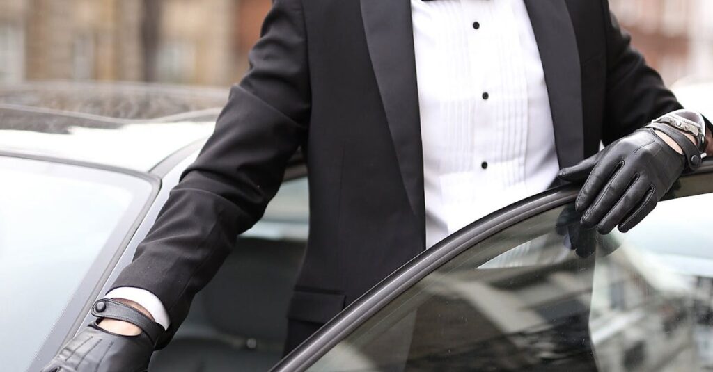 Well-dressed man in a tuxedo standing confidently by his car in an urban setting.