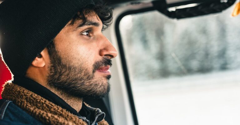 Man with a beard in a warm jacket driving through a snowy landscape, showcasing adventure and travel.