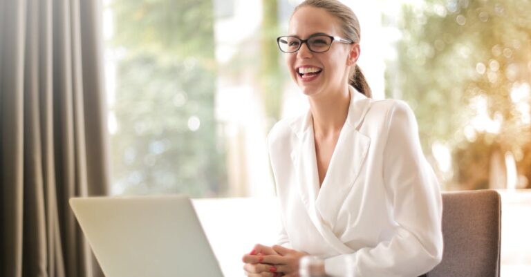 Cheerful businesswoman in glasses working on a laptop, in a bright and modern office setting.