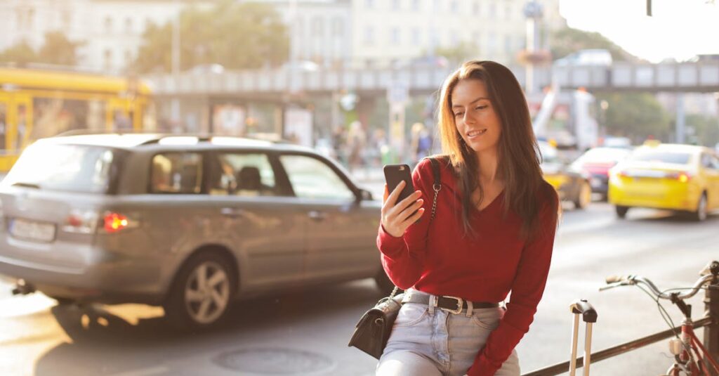 Smiling woman in a red sweater uses her phone on a sunny urban street. Cars and bicycles surround her.