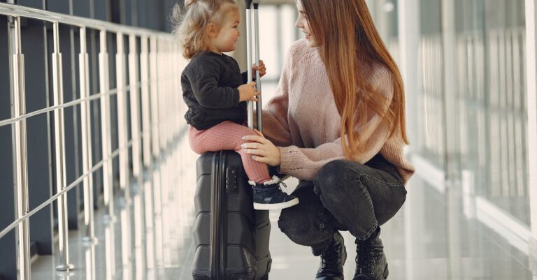 Full length of positive content woman and cute stylish blond little girl sitting on black suitcase in airport corridor while waiting for flight