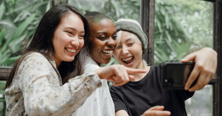 Three friends of different ethnicities laughing and taking a selfie together indoors.