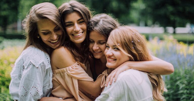 Four smiling women joyfully embrace in a lush green park during summer, showcasing genuine friendship and happiness.