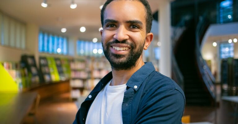 Smiling ethnic bearded student with toothy smile standing against bookshelves in library in college