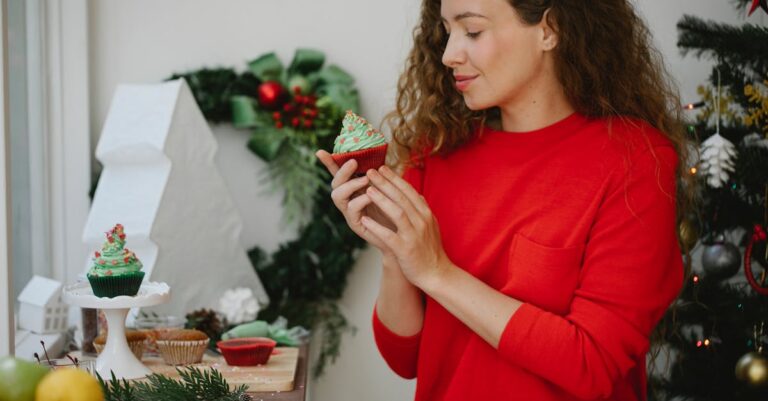 Happy young woman enjoying fresh delicious cupcake with green whipped cream standing beside decorated Christmas fir tree