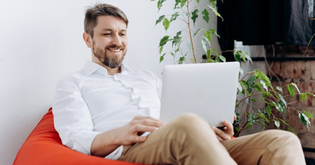 Man sitting on red bean bag chair using laptop, smiling in casual modern workspace.