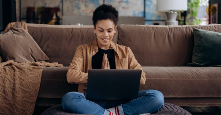 A smiling young woman sits cross-legged on a pouf, using her laptop in a cozy home setting.