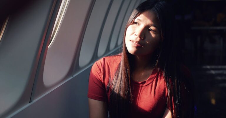 A woman in a red shirt sits in an airplane, gazing thoughtfully out the window as sunlight illuminates her face.