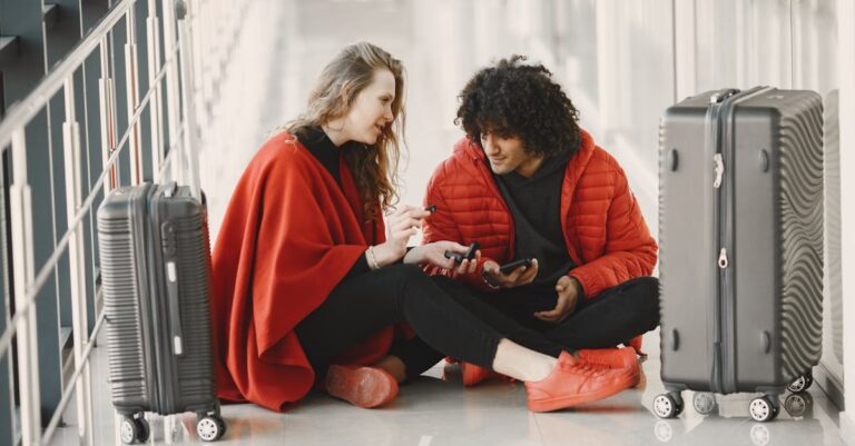 A young couple sits on the floor at an airport, engaging and relaxed, with suitcases around them.