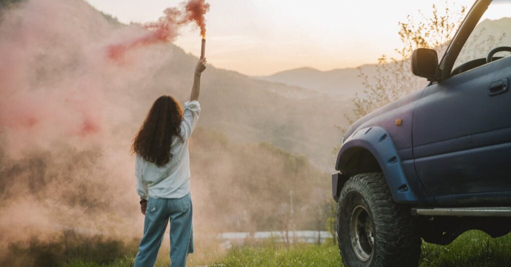 Woman with smoke flare and SUV in scenic mountain landscape at sunset.