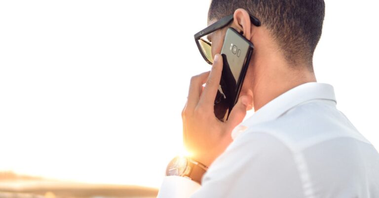 Man outdoors in Morocco talking on smartphone, wearing sunglasses and white shirt, backlit by sunset.