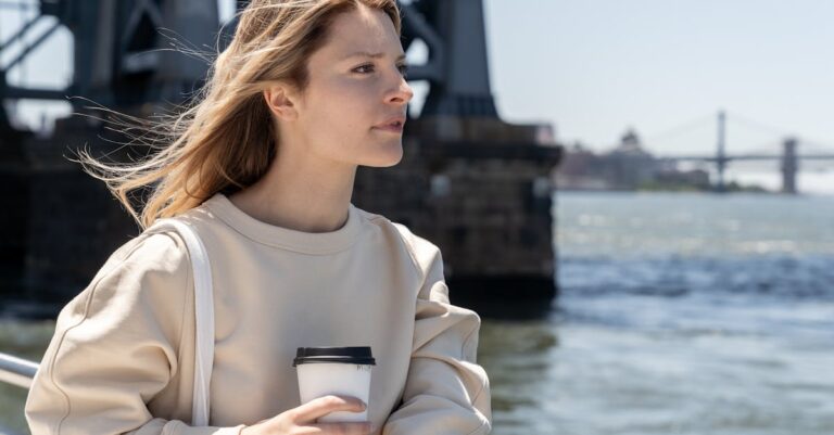 A young woman enjoys a coffee by a scenic riverside with a bridge view.