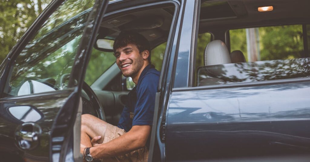 A cheerful man sits inside a car with the door open, enjoying a sunny day outdoors.