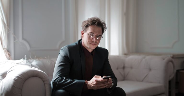 Businessman in a black suit sitting on a sofa, using a smartphone. Bright and modern indoors setting.