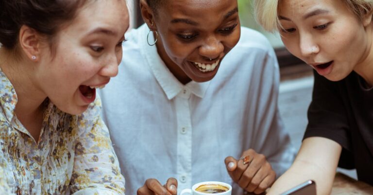 Amazed surprized multiethnic female friends in casual clothes with cup of coffee browsing smartphone while sitting at wooden table with laptop against window