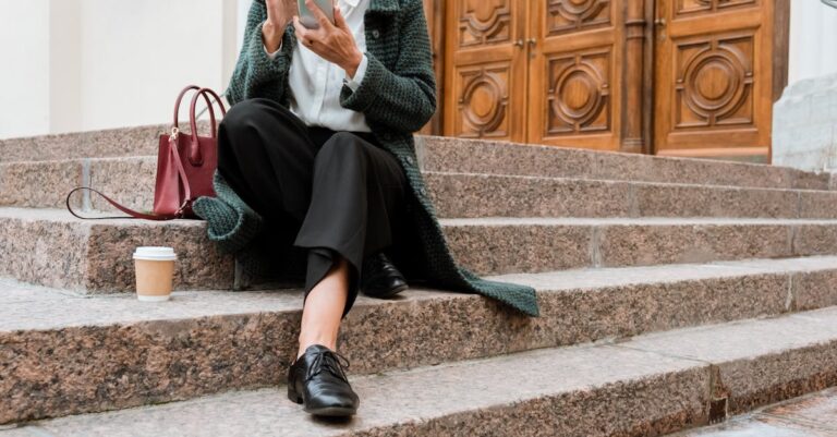 Woman sits on stone steps using smartphone with coffee and handbag, outdoors near wooden doors.