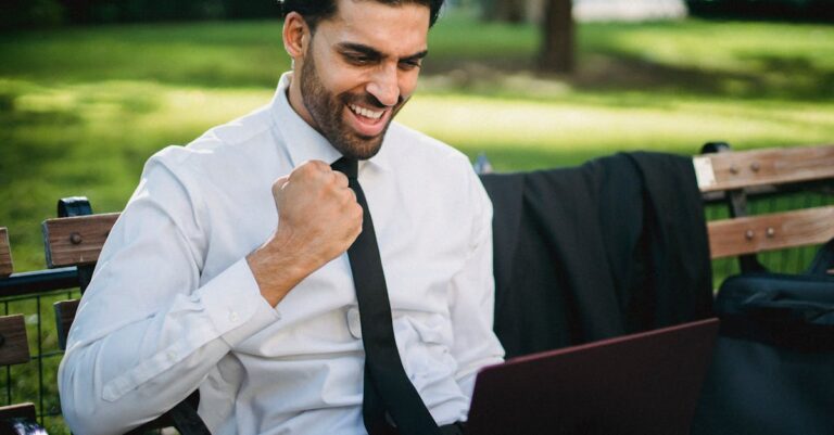 A joyful businessman celebrates a success working on his laptop outdoors on a park bench.