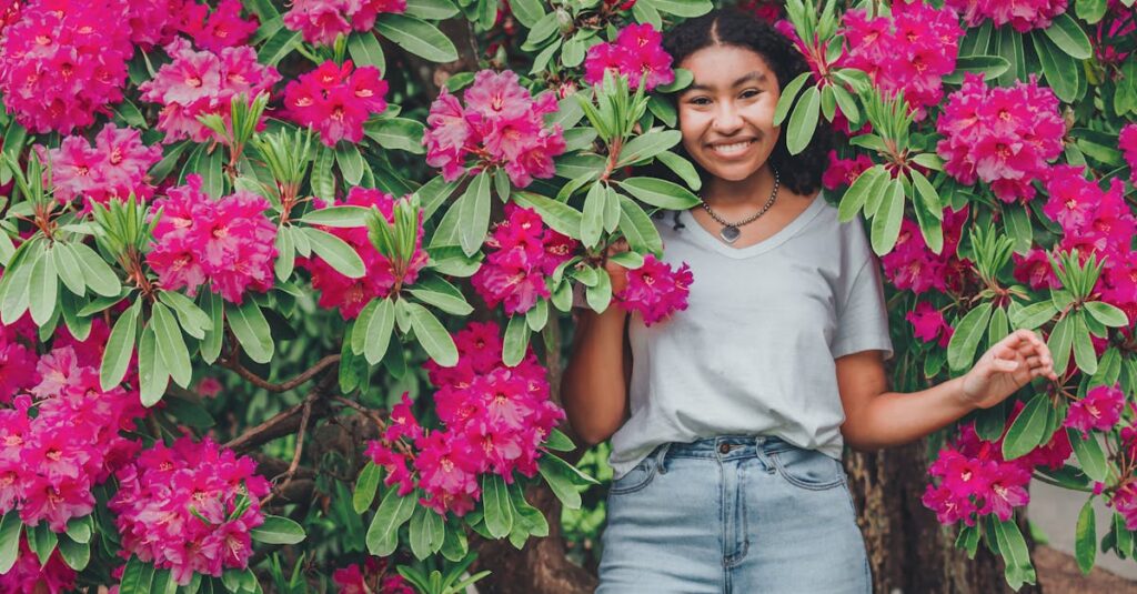 A cheerful young woman smiles amidst vibrant pink rhododendron blooms in a lush spring setting.