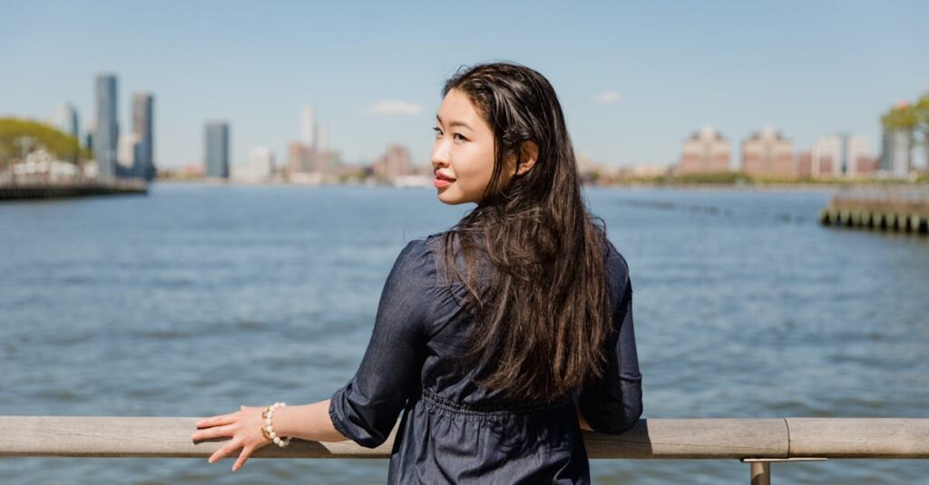 Asian woman gazes at city skyline from river railing, creating a serene moment outdoors.