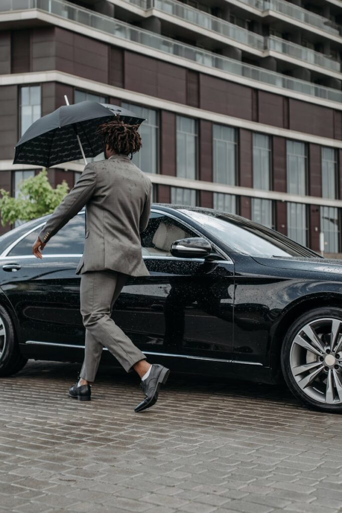 Man holding umbrella opens car door on rainy day in front of modern building.