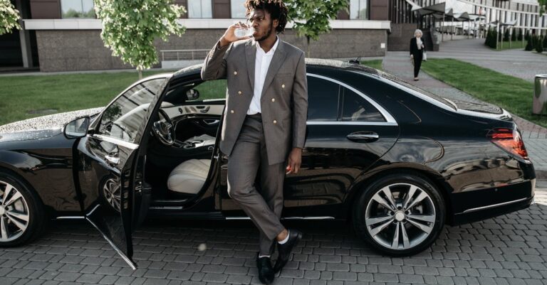 A fashionable businessman stands by a luxury car, enjoying a drink on a city street.