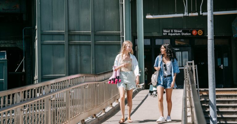 Two young women casually walking on a footbridge outside NY Aquarium Station on a sunny day.