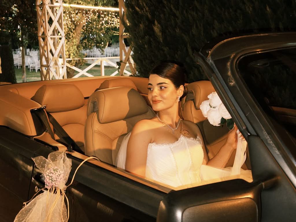 Bride in a convertible car at a night wedding, holding a bouquet. Captures elegance and celebration.