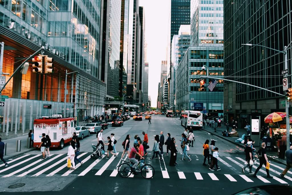 Vibrant New York City street with diverse pedestrians crossing at dusk, framed by towering skyscrapers.